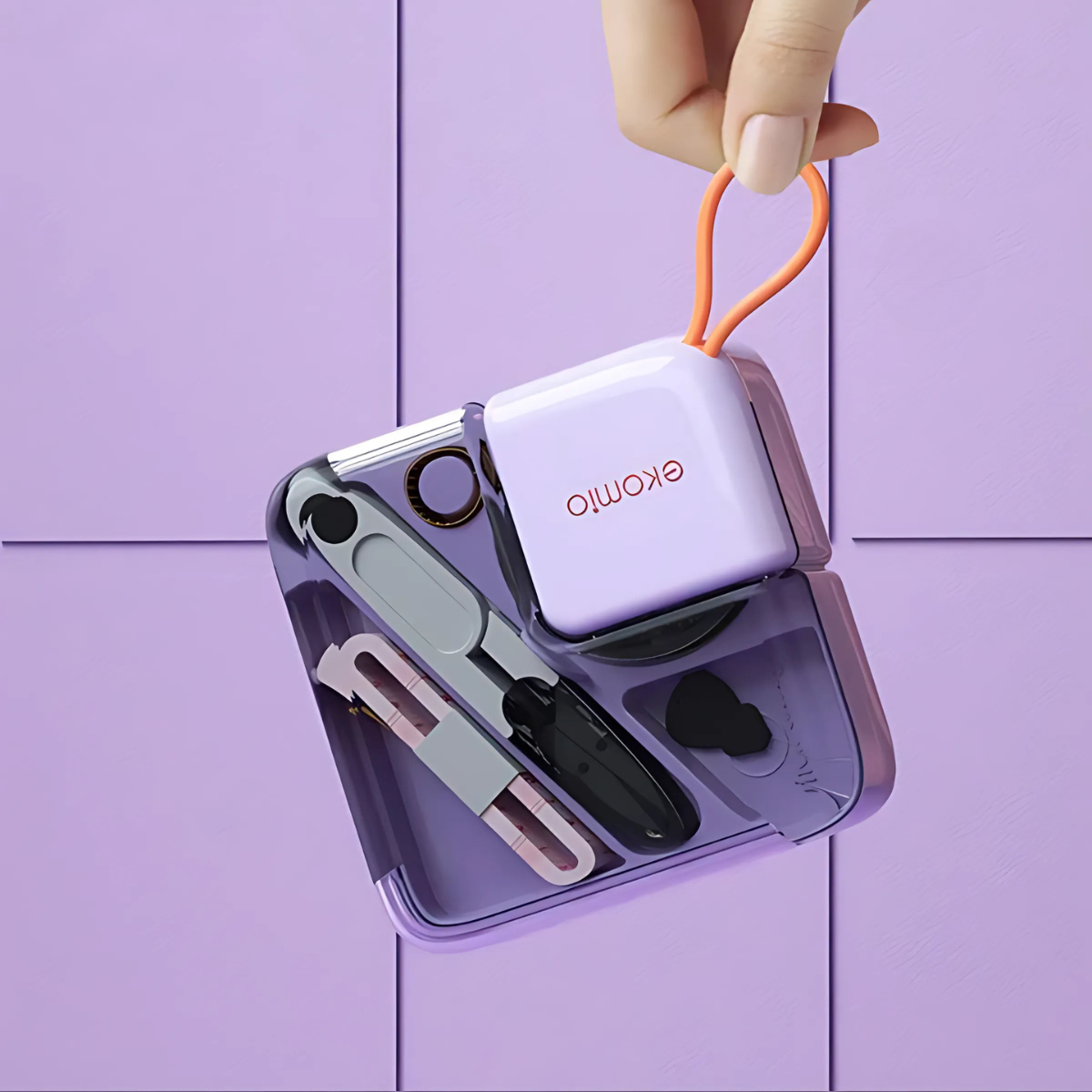 A hand holds a compact purple and orange Mini Sewing Kit labeled "ekomo" next to an open purple container displaying various sewing items, all arranged on a purple tiled surface.