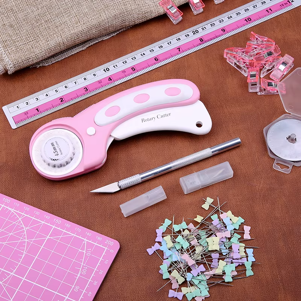 The 39-Piece Quilting Kit: Perfect for Beginners includes essentials like a pink rotary cutter, acrylic ruler, craft knife, sewing clips, cutting mat, colorful pins, and fabric—all arranged on a brown surface.