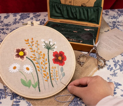 A person holds an embroidery hoop with colorful flowers while a compact wooden case from the Vintage Antique Sewing Kit, featuring an antique bronze finish and vintage scissors, sits on a floral tablecloth.