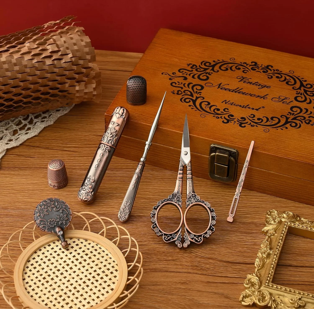 The Vintage Antique Sewing Kit sits on a wooden table, featuring scissors, thimbles, and a needle case all in a compact wooden case. An antique bronze finish enhances its timeless charm.