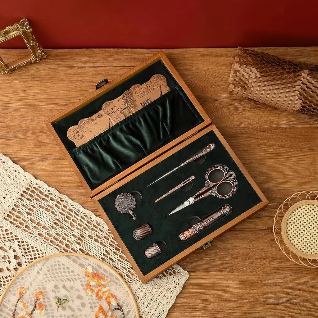 The Vintage Antique Sewing Kit comes in a compact wooden case with scissors, needles, and thimbles elegantly arranged on dark green fabric. Nearby, lace fabric and an embroidery ring rest on the surface, showcasing its timeless charm.